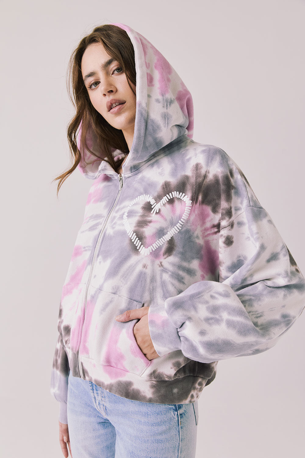 Side view of woman wearing Chaser’s Heart Tie Dye Zip Up Hoodie with heart embroidery detail and hood up on a white background