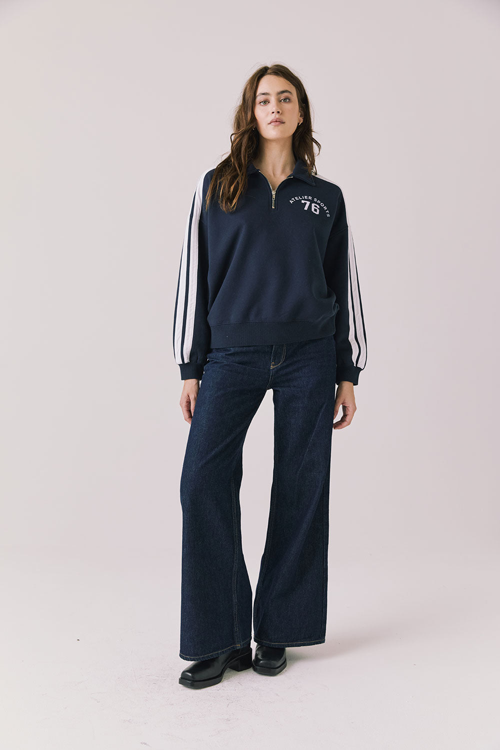Woman wearing Chaser's navy Atelier Sports 76 Half Zip Long Sleeve with white stripes and dark denim jeans on a light gray background
