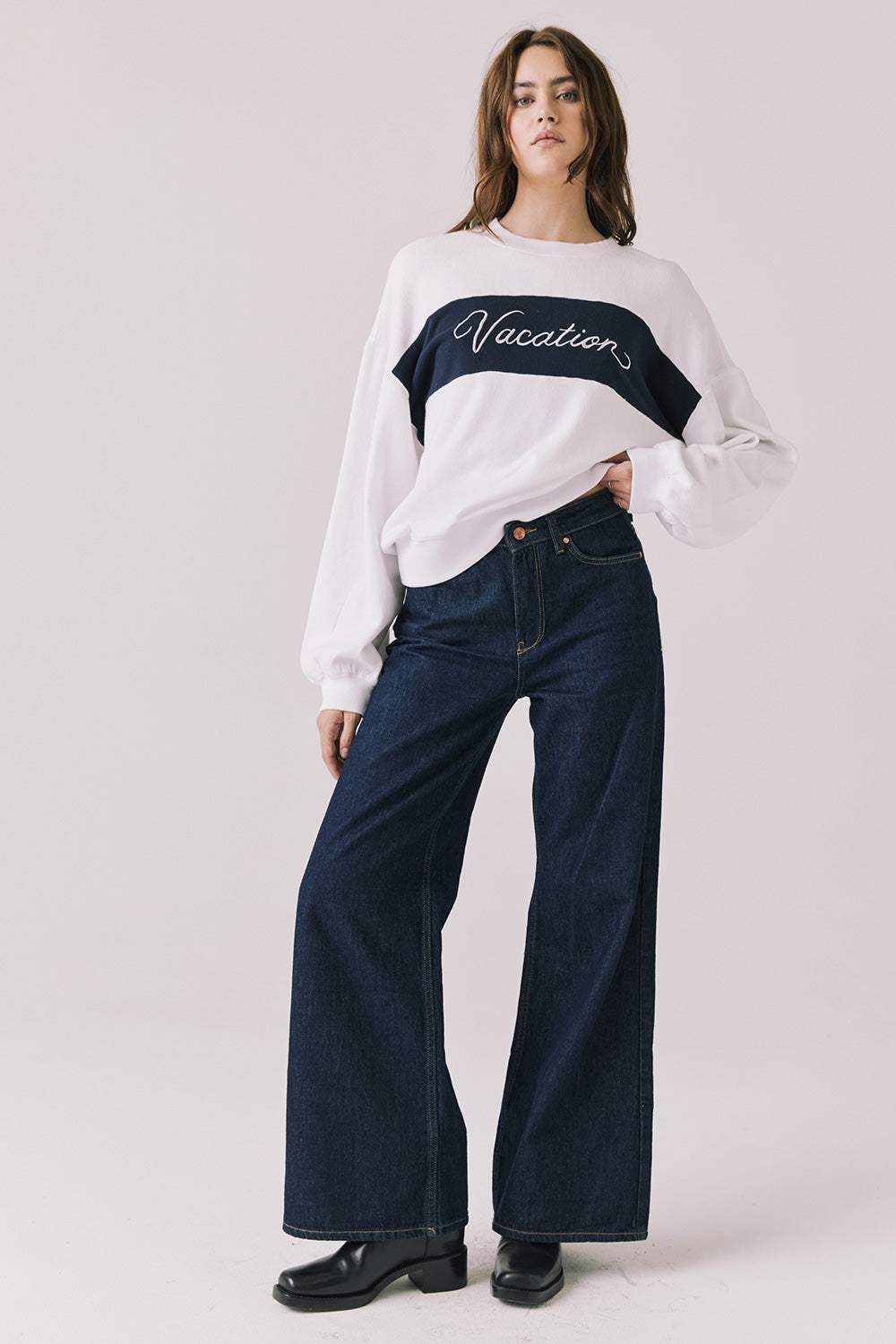Full body view of woman wearing Chaser’s white Vacation Long Sleeve with ‘Vacation’ text and dark jeans on a light background