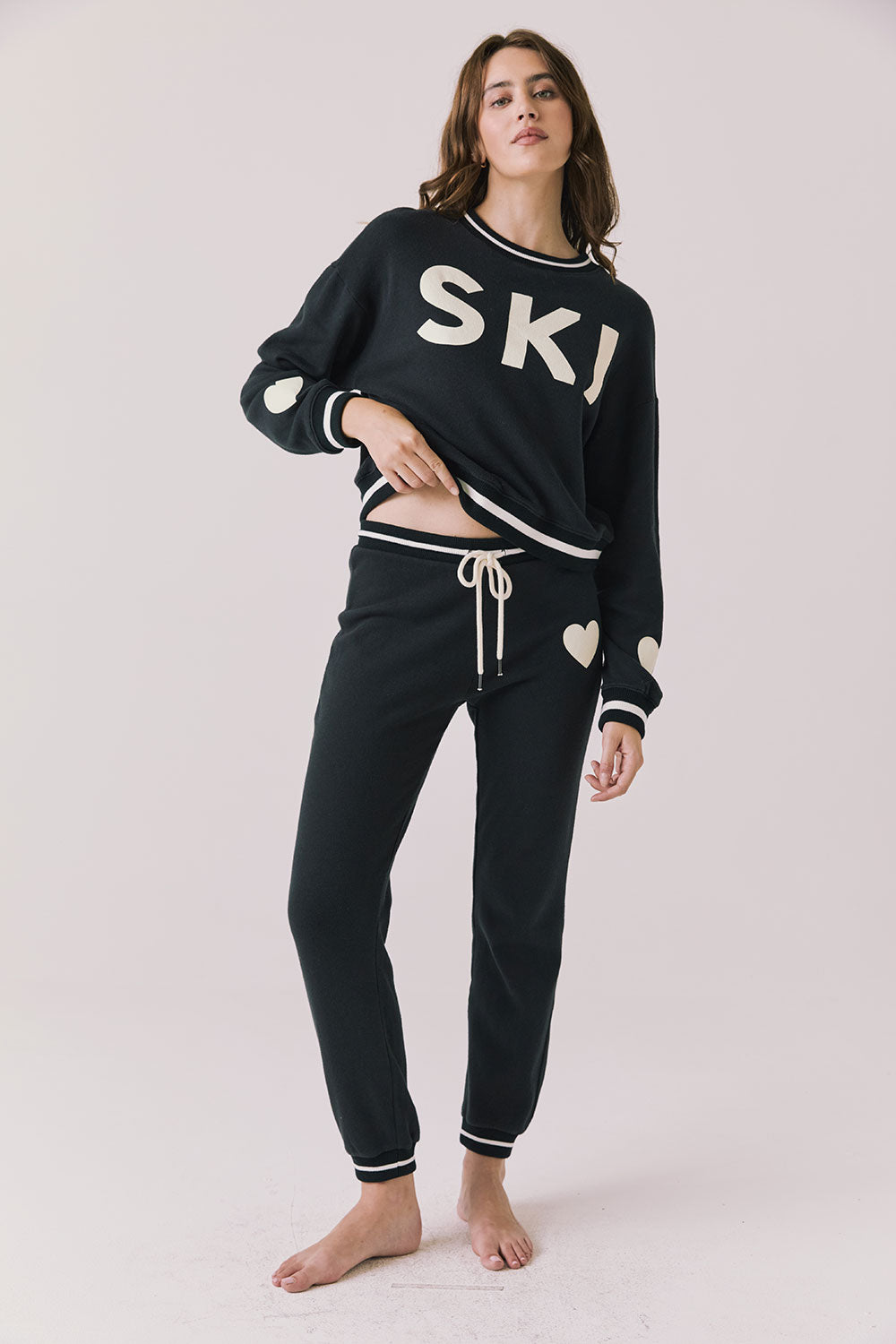 Full body view of woman wearing Chaser’s Heart True Black Joggers with matching “SKI” pullover on a white background
