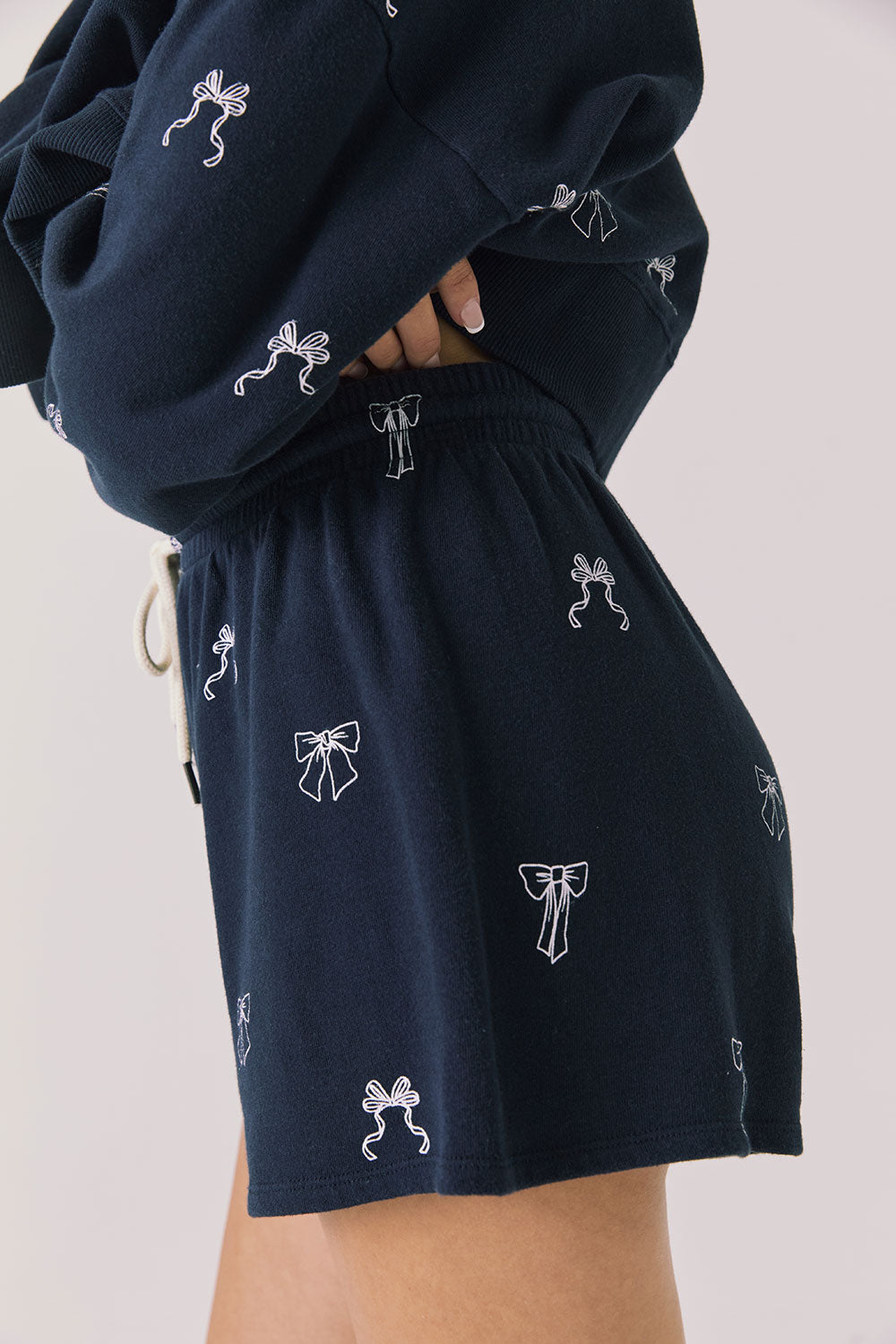 Side view of woman wearing Chaser’s navy Tossed Bows Shorts with allover bow pattern on a white background