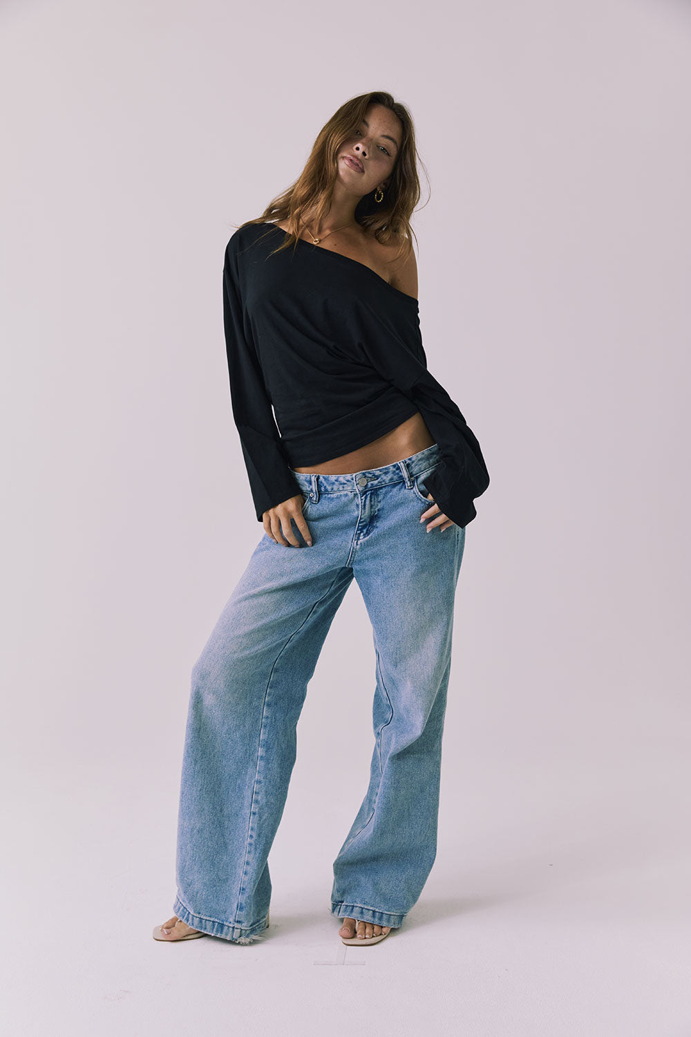Full body view of a woman wearing Chaser off-shoulder Ash Black Long Sleeve and blue jeans against a plain background