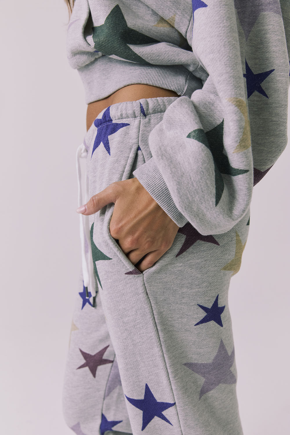 Close up side view of the Chaser You’re A Star Pants in Heather Grey showing the star print soft fleece fabric with hand in pocket and elastic waistband detail