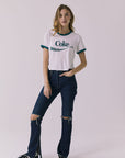 Full body front view of a woman wearing Chaser's white Coca Cola The Real Thing tee with 'Coke' logo and blue jeans on a plain background