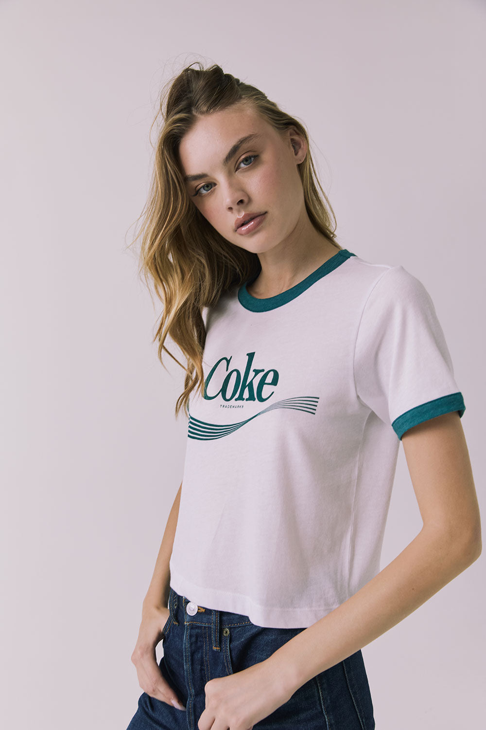 3/4 view of a wearing Chaser's white Coca Cola The Real Thing tee with 'Coke' logo and blue jeans on a plain background