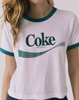 Close up front view of a woman wearing Chaser's white Coca Cola The Real Thing tee with 'Coke' logo and blue jeans on a plain background
