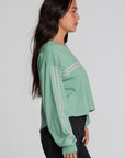 A woman in profile wearing a sage green cropped long-sleeve sweatshirt with white double stripes on the sleeves, paired with black jeans, against a light gray studio background