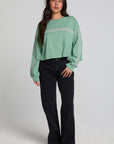 A woman modeling a sage green long-sleeve crewneck sweatshirt with thin white horizontal stripes across the chest and sleeves, paired with black wide-leg jeans and brown shoes, against a light gray studio background