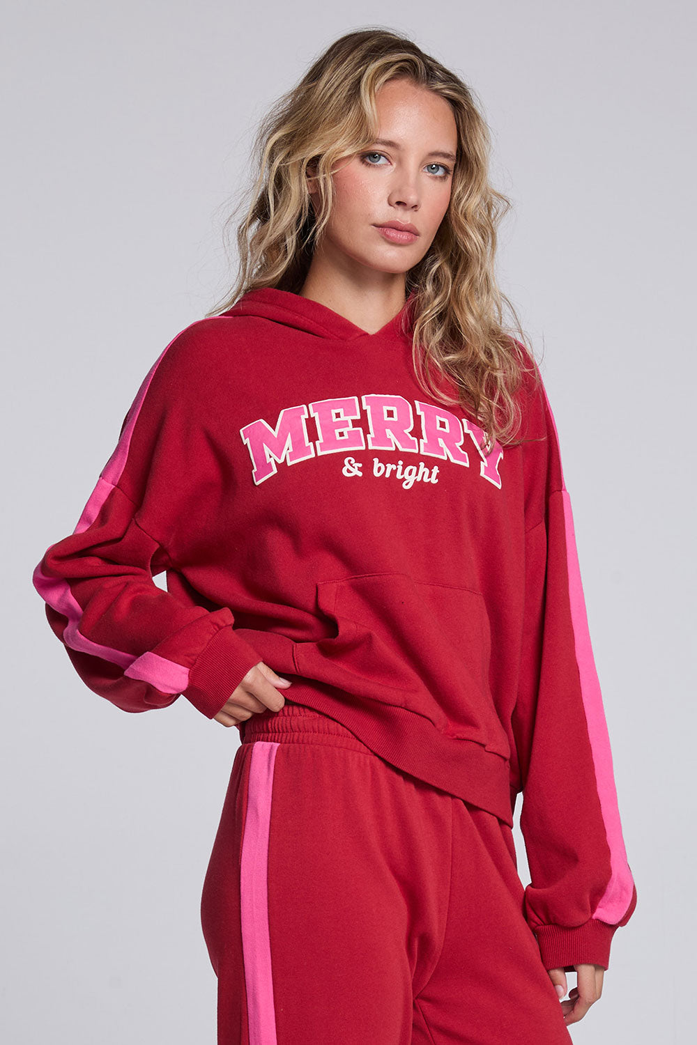 Model wearing Chaser's Merry and Bright Hoodie in Cranberry Velvet with varsity-style Merry and Bright artwork, angled view