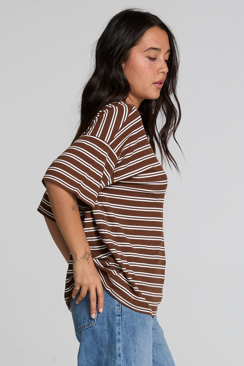Side view of a woman wearing the Chaser Peri Nutty Brown Stripe Tee and blue jeans on a neutral background