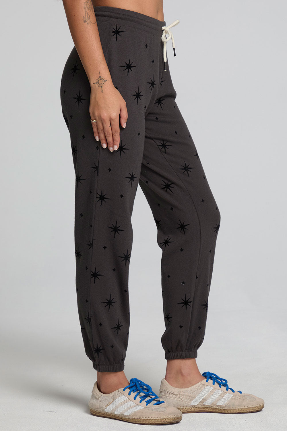 Side view of a woman wearing the Chaser  Allover Mini Stars Pants on a neutral background with hands at sides