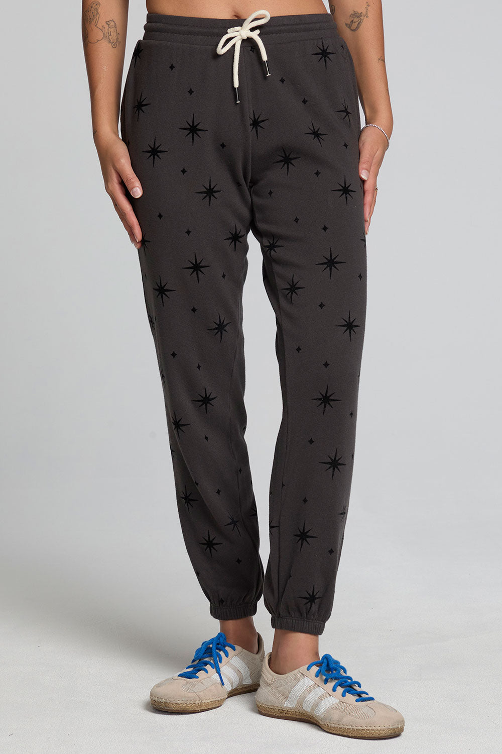 Front view of a woman wearing the Chaser  Allover Mini Stars Pants on a neutral background with hands at sides and sneakers