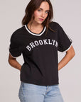Front view of a woman wearing the Chaser Brooklyn Varsity Graphic Tee on a neutral background with hands on hips
