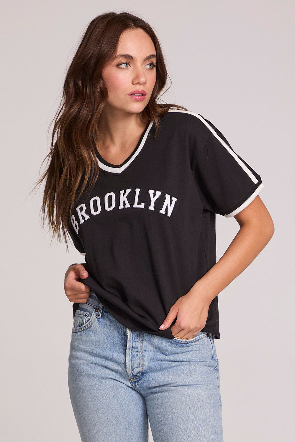 Front view of a woman wearing the Chaser Brooklyn Varsity Graphic Tee on a neutral background with one hand in her pocket