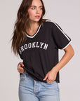 Front view of a woman wearing the Chaser Brooklyn Varsity Graphic Tee on a neutral background with one hand in her pocket