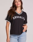 Front view of a woman wearing the Chaser Brooklyn Varsity Graphic Tee on a neutral background with one hand on hip