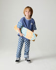 Front view of a boy wearing the Chaser Smileys Boys Tee on a neutral background with matching checkered pants holding long board