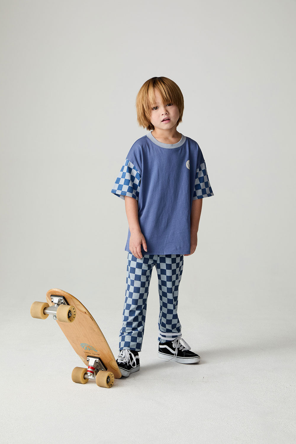 Front view of a boy wearing the Chaser Smileys Boys Tee on a neutral background with matching checkered pants stepping on long board