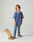 Front view of a boy wearing the Chaser Smileys Boys Tee on a neutral background with matching checkered pants stepping on long board
