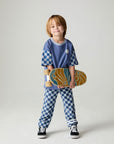 Front view of a boy wearing the Chaser Smileys Boys Tee on a neutral background with matching checkered pants holding long board while smiling