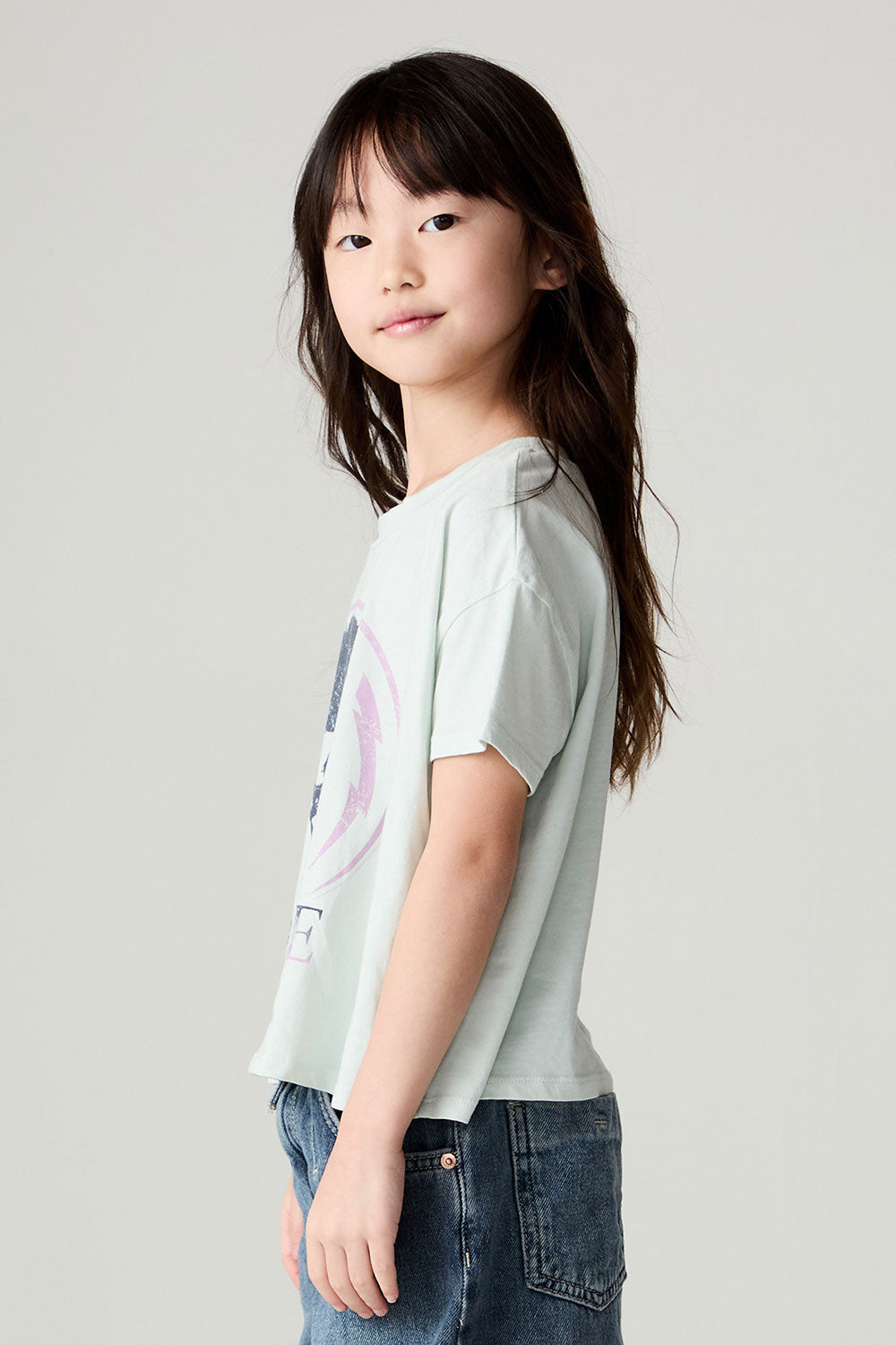 Side view of a girl wearing the Chaser David Bowie Stars Bowie Girls Tee on a neutral background with bluejeans