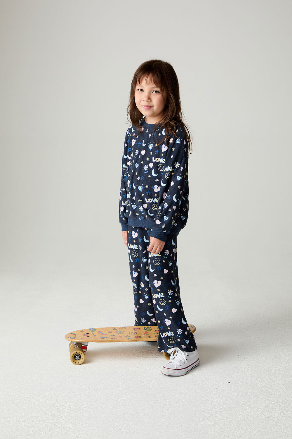 Front view of a girl wearing the Chaser Free Draw Girls Tee and matching pants on a neutral background posing with a skateboard