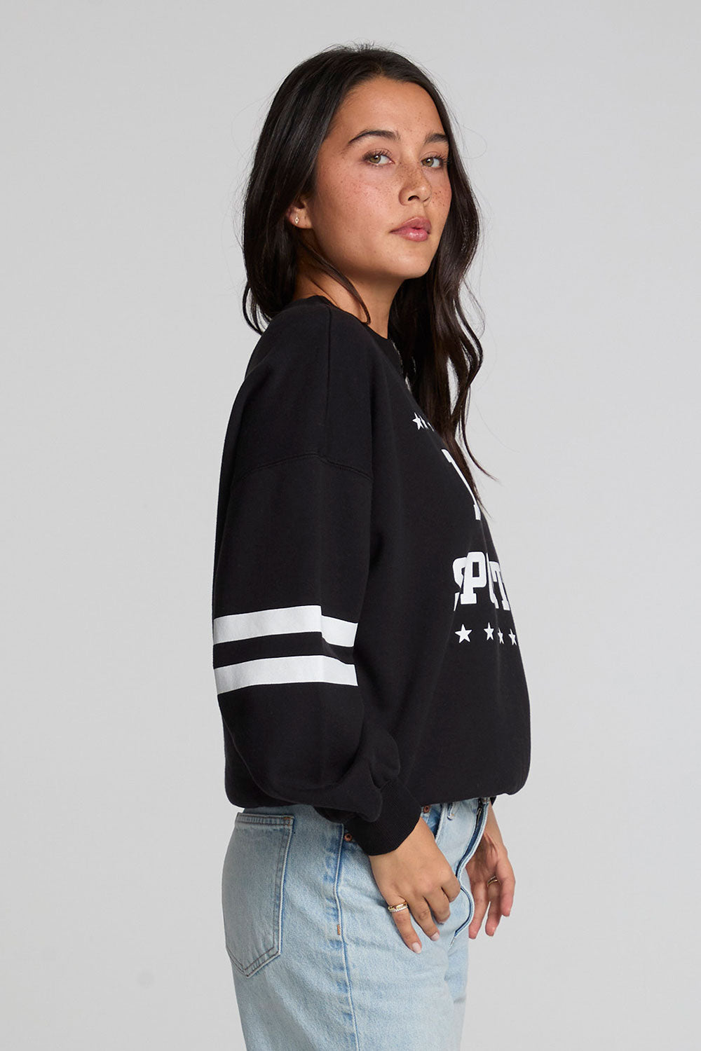 Side view of a woman wearing the Chaser I Love Sports Sweatshirt on a neutral background with jeans and hands on pockets