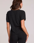 Back view of a woman wearing the Chaser   Leonor Black Tee on a neutral background with black pants and hands down