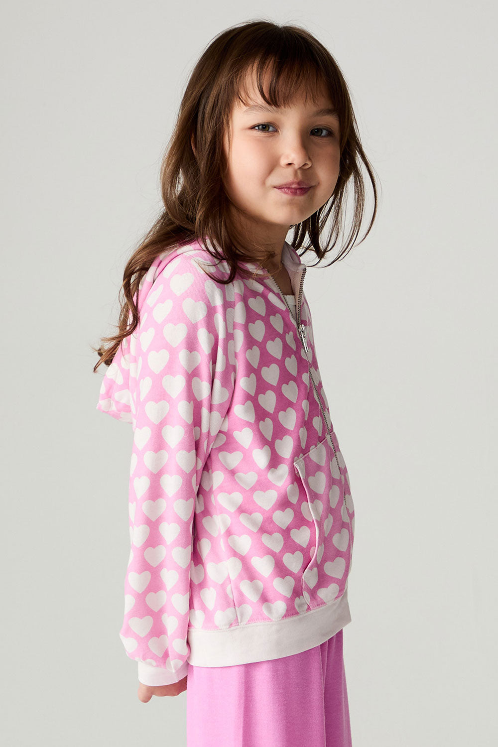 Side view of a girl wearing the Chaser  Love Is Everywhere Girls Hoodie and matching pink pants with arms lowered