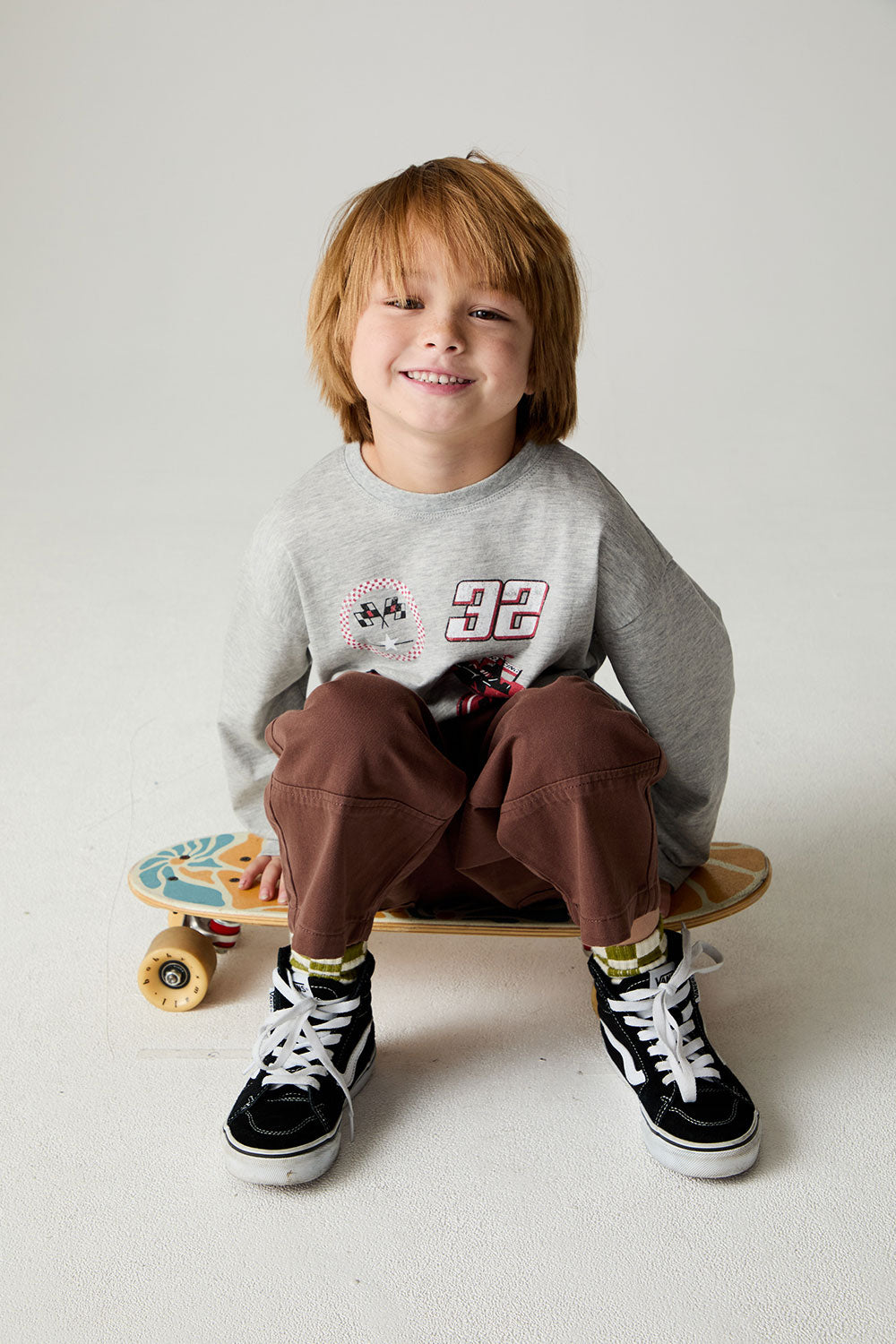 Seated boy on skateboard wearing the Chaser Moto Patches Boys Tee on a neutral background 
