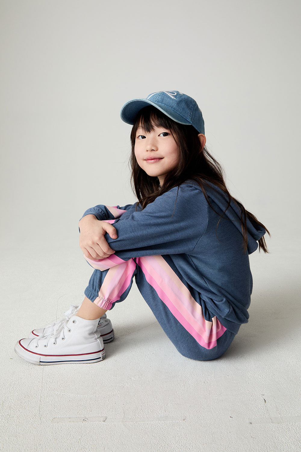 Front view of a seated girl wearing the Chaser Multi Stripes Girls Hoodie on a neutral background with hat and matching pants