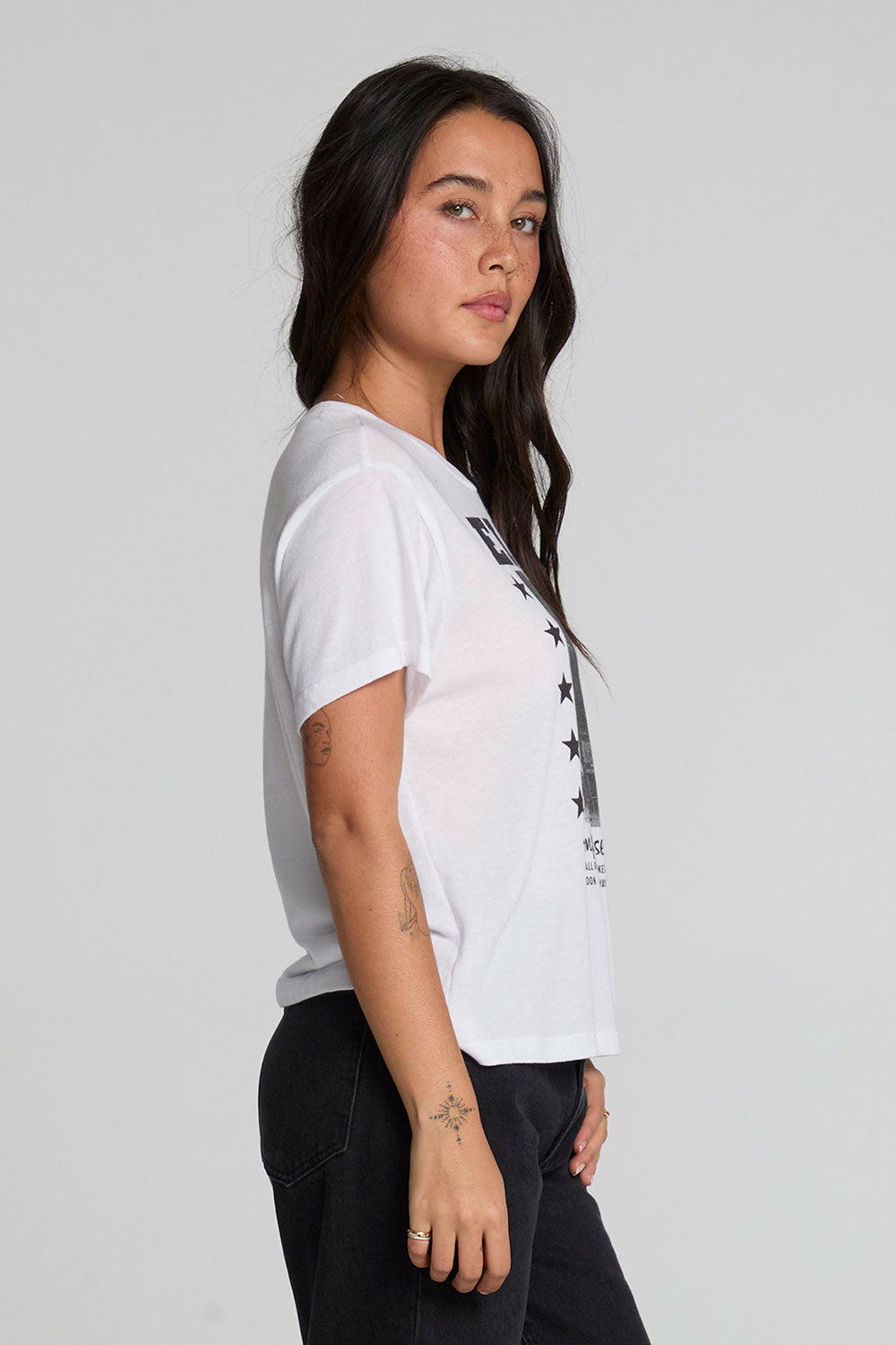 Side view of a woman wearing the Chaser Sun Records Elvis 1972 Tee on a neutral background with arms at sides and pants