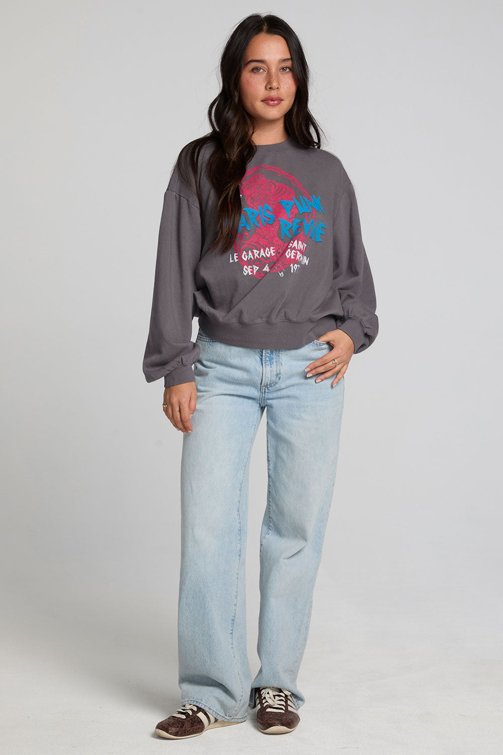 Full body view of a woman wearing the Chaser The Paris Punk Graphic Sweatshirt and jeans on a neutral background with arms lowered and one hand on hip