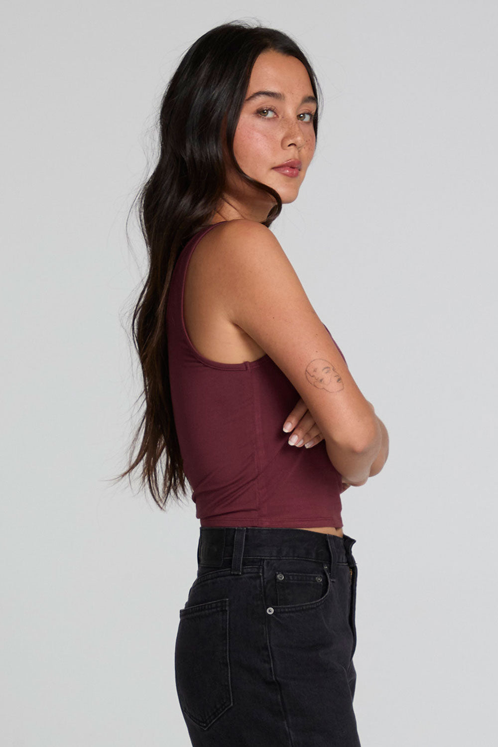 Side view of a woman wearing the Chaser Verze Brick Tank and jeans on a neutral background with arms crossed 