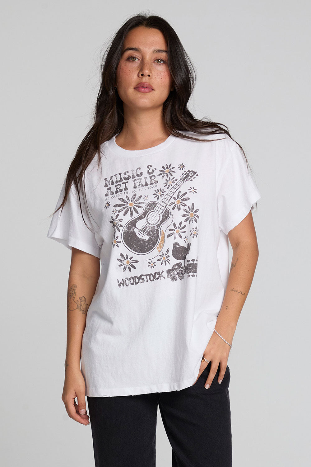 Front iew of a woman wearing the Chaser Woodstock Music And Art Fair Tee on a neutral background with black pants and hand on one pocket