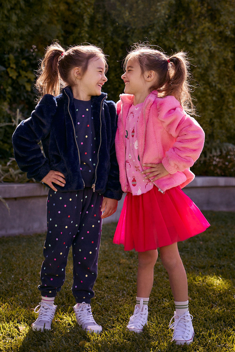 Chaser Kids Avalon Girls Puff-Sleeve Jacket in navy and Camelia Girls Puff-Sleeve Jacket in pink, worn by two girls on grass beside a low wall and greenery