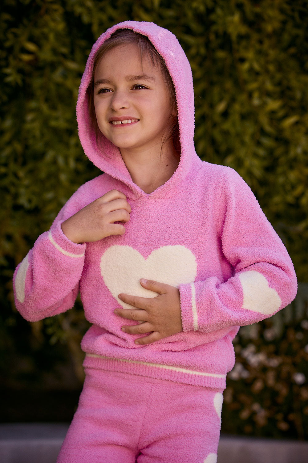 Chaser Kids Holiday Hug Girls Hoodie in pink with white hearts, close-up with hood up outdoors in front of greenery