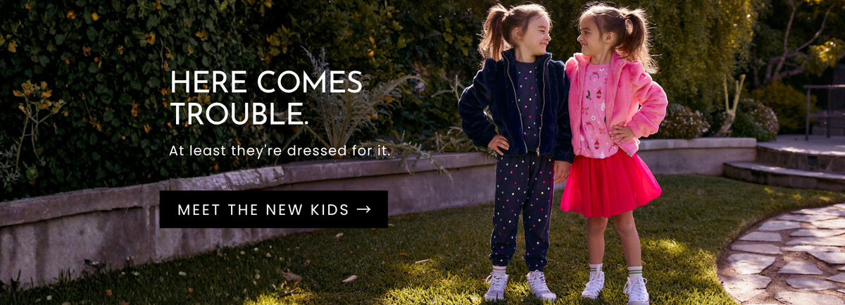 Two girls wearing Chaser Kids Holiday 2026 outfits, one in a navy polka dot matching set with a fuzzy jacket and the other in a pink graphic top, fuzzy jacket and bright pink skirt, smiling at each other outdoors