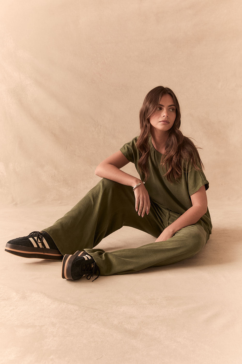 Full front view of women sitting wearing Chaser's Lee Trouser in bronze green with wide-leg silhouette and relaxed drape with matching tee against a beige background