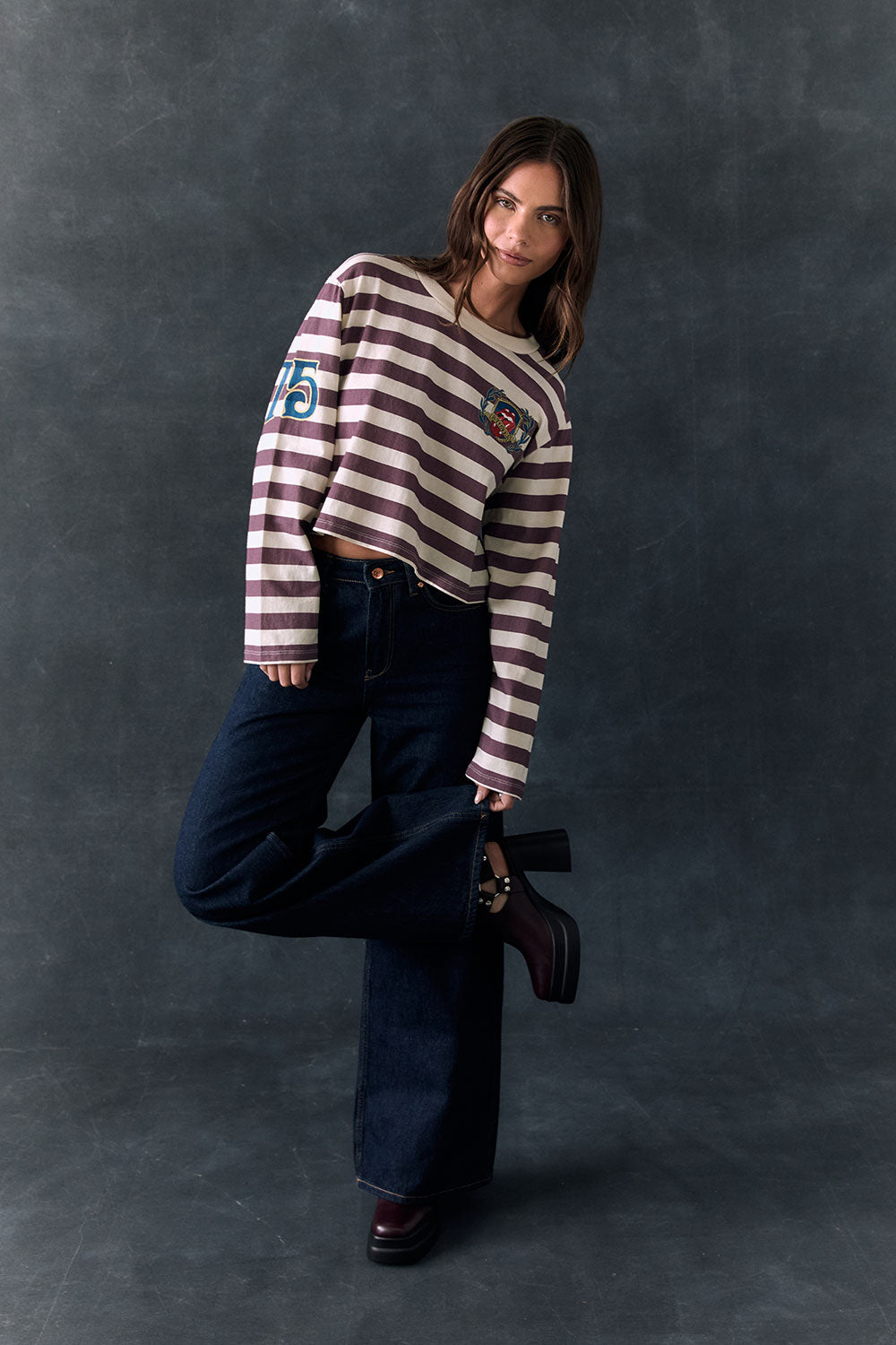 Full body view of model standing against a dark background wearing The Rolling Stones Rugby Stones Logo Long Sleeve with red and white stripes, cropped fit, tongue logo graphic, and jeans

