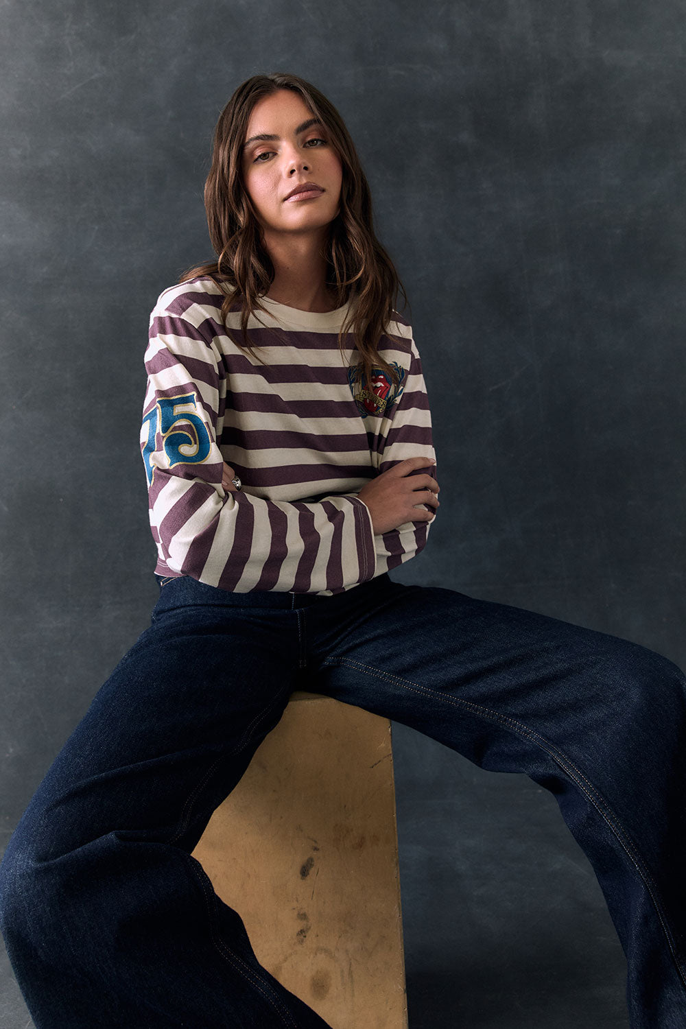 Full body view of model sitting on a wooden block against a dark background wearing The Rolling Stones Rugby Stones Logo Long Sleeve with red and white stripes, cropped fit, tongue logo graphic, and jeans
