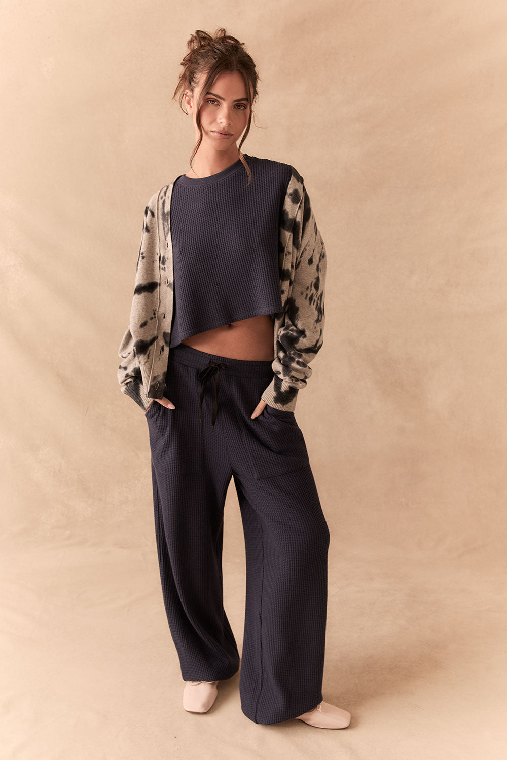 Full body view of Woman wearing a patterned cardigan over a crop top and wide-leg pants on a beige background