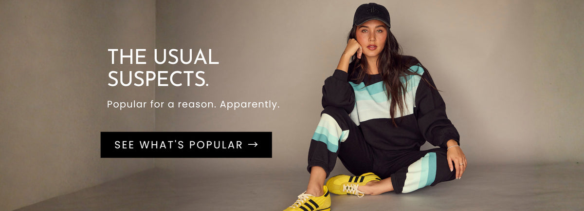 Model seated in Chaser's Cashbah Black Pullover and matching Ada Black Jogger with turquoise and white colorblock stripes, styled with bright yellow sneakers for the Best Sellers collection